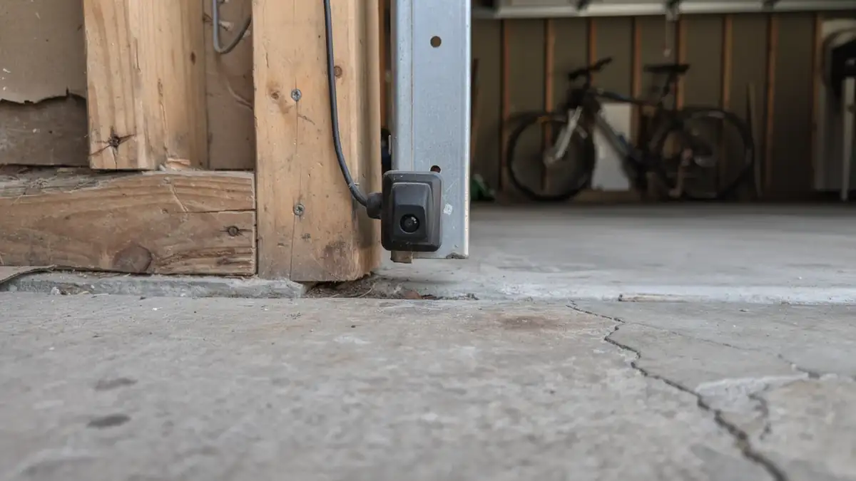 A garage door safety sensor is mounted on a wooden frame next to a concrete floor.